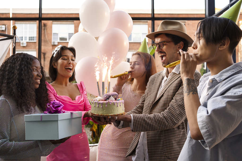 Festa na empresa com um grupo de cinco funcionários felizes festejando, uma deles está segurando um presente e um deles segura um bolo com velas acessas