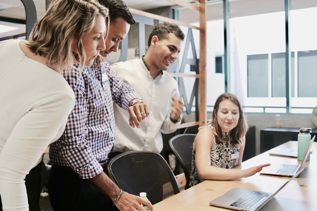 Grupo de quatro pessoas em sala de reunião, uma mulher mostra para os colegas algo na tela de um notebook e eles estão sorrindo, atentos à tela, simbolizando retenção de talentos