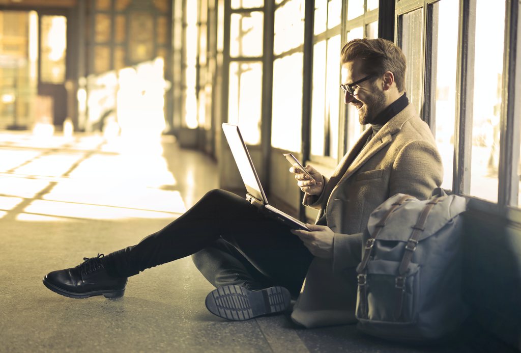 Colaborador sentado no chão de aeroporto, sorrindo usando celular, com notebook no colo e mochila ao lado. Simbolizando ajuda de custo no cartão pré-pago empresarial