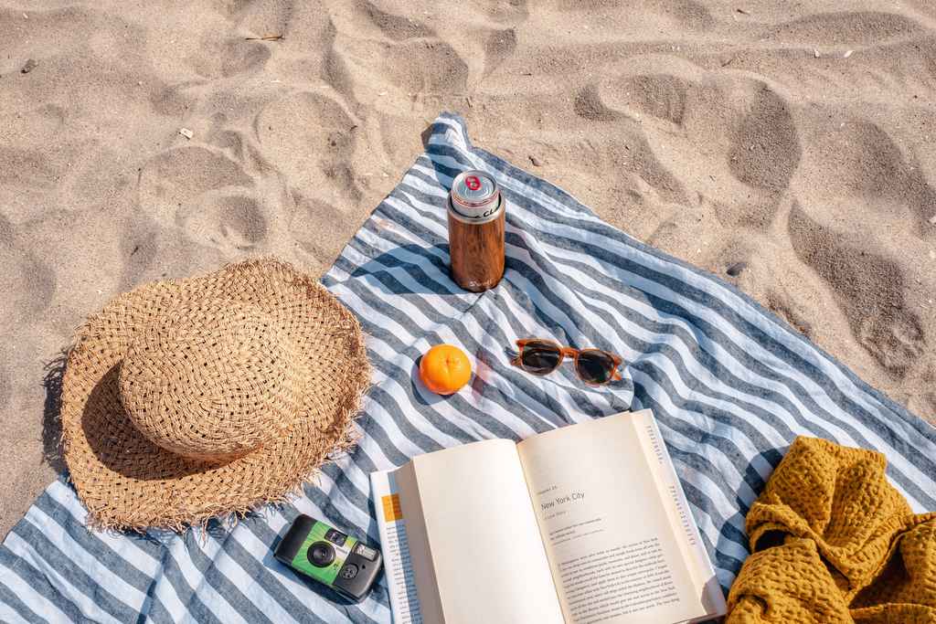 Areia de praia com pano de piquenique para proteger uma latinha de refrigerante, um a tangerina, um óculos de sol, um chapéu de palha, uma câmera fotográfica e um livro aberto. A foto simboliza Retorno de ferias ao trabalho